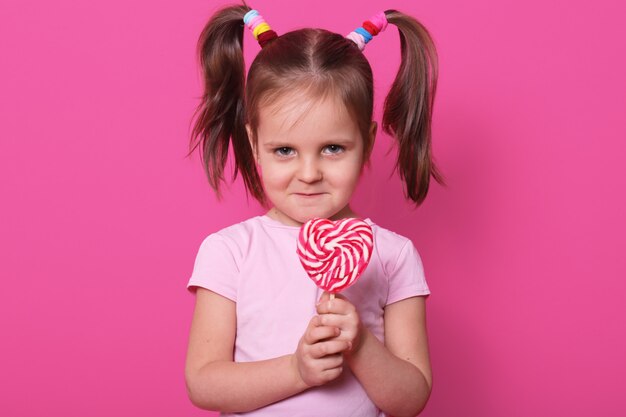 fair haired child stands straight, holds colourful lollipop in hands, having frowny face, has no desire to share candy with anybody. Copy space for advertisement.