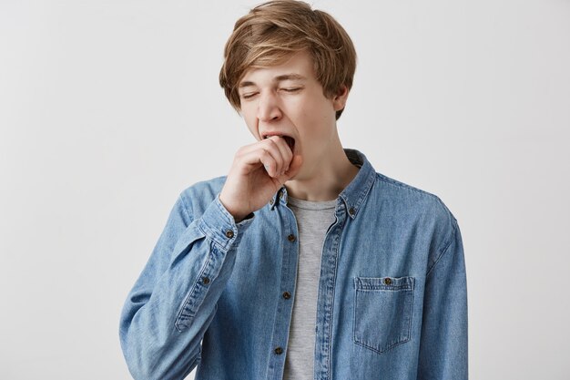 Fair-haired guy in denim shirt looking tired and sleepy, yawning, covering mouth with, after sleepless night. Male student looking bored and disinterested during classes at university. Body language
