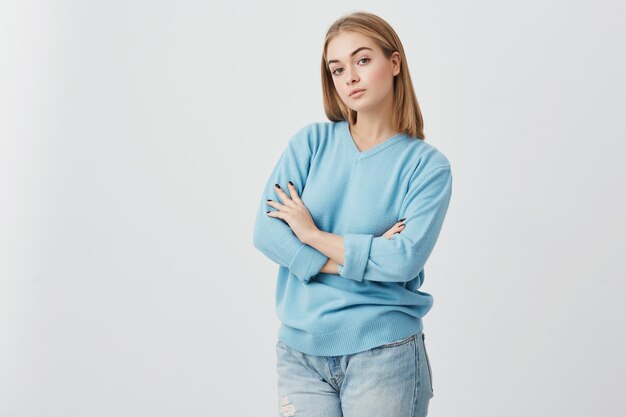 Fair-haired woman with warm dark eyes and healthy skin, standing with folded arms, looking directly into camera. Alluring girl with beautiful appearance dressed casually posing in studio.