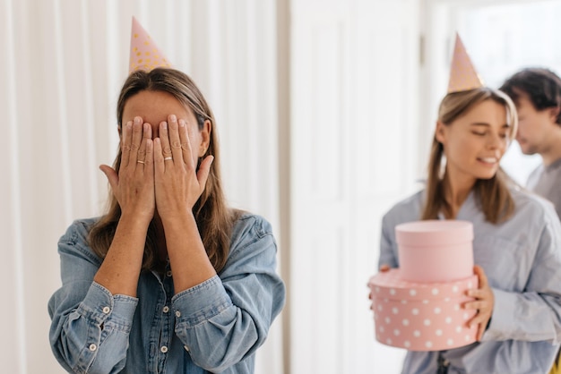 Fair-skinned woman covers her face with hands in anticipation of long-awaited gift from friend indoors. celebration concept