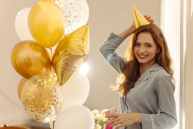 Fairskinned young redhaired girl wears nightgown festive cap standing near balloons with glass Youth holiday concept