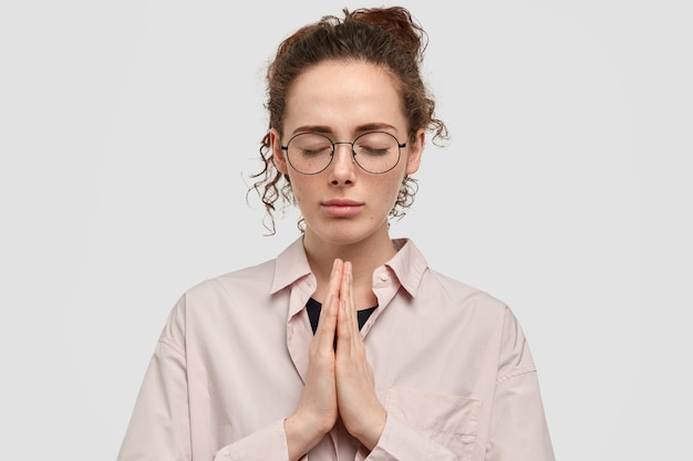 Бесплатное фото Верная веснушчатая женщина держит руки в молитвенном жесте, верит в чудо, закрывает глаза,