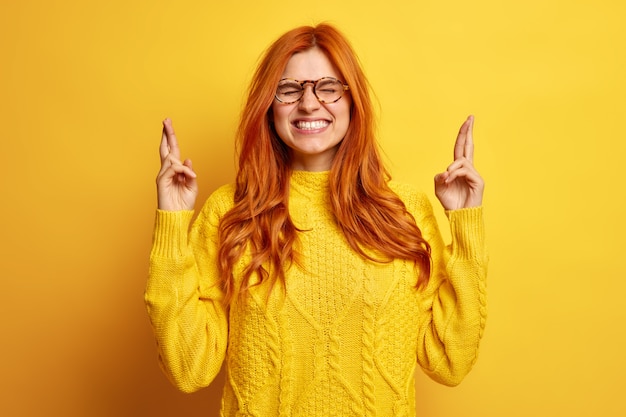 Free photo faithful pretty young redhead woman closes eyes keeps fingers crossed makes wish dressed in casual jumper believes in good luck.