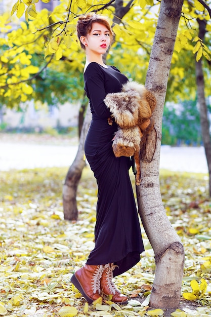 Fall fashion lifestyle portrait of sexy beautiful asian woman wearing long stylish maxi dress boots and holding piece of fur, posing at city park in nice sunny autumn day. Bright colors.