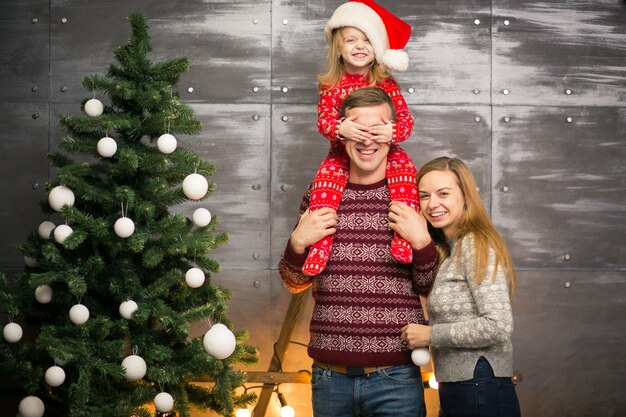 Family by the Christmas tree with little daughter in a red hat