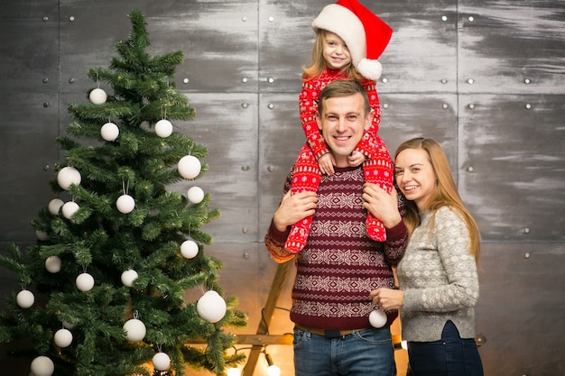 Family by the Christmas tree with little daughter in a red hat