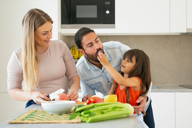 Family cooking and eating at home during pandemic. Girl giving slice of veg to dad for taste while mom cutting fresh vegs and fruits. Family cooking or lifestyle concept