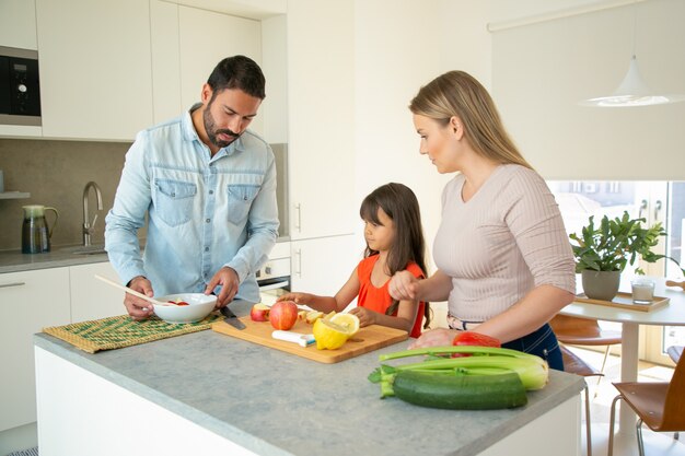 Family cooking home dinner during pandemic. Young couple and kid cutting vegetables for salad at kitchen table. Healthy nutrition or eating at home concept