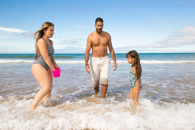 Family couple and little girl walking ankle deep in sea water, picking shells into bucket
