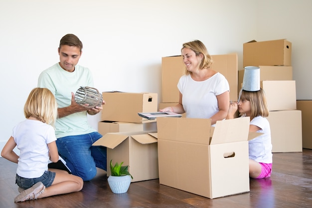 Family couple and little girls moving into new flat, having fun while unpacking things in new apartment, sitting on floor and taking objects from open boxes