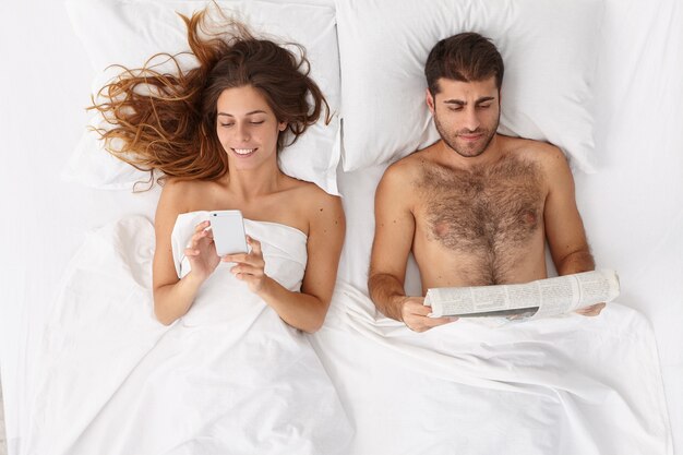 Family couple stay in comfortable bed before sleep, woman uses mobile phone for online chatting, surfs internet, addicted to moden technologies, man reads newspaper, do not talk to each other