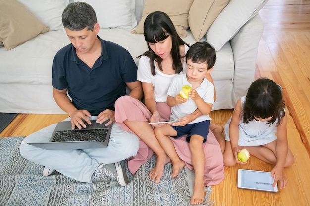 Family enjoying leisure time together, using digital gadgets and eating fresh apples in apartment.