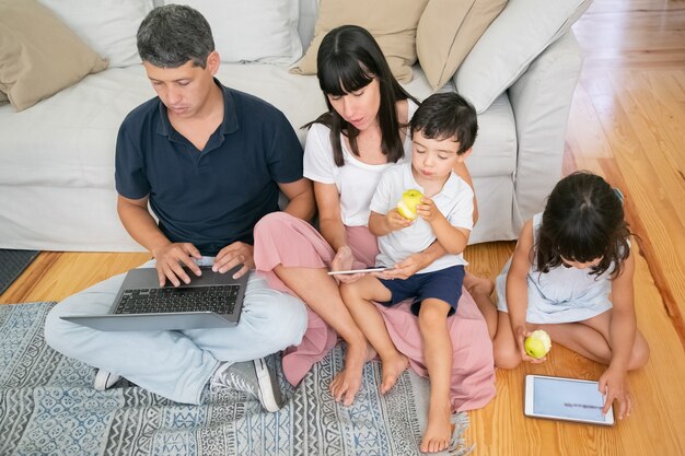 Family enjoying leisure time together, using digital gadgets and eating fresh apples in apartment.
