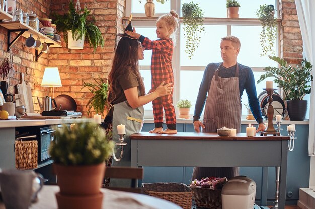 Family in halloween. Little girl standing on table and dresses a magic hat on her mom head in loft style kitchen at morning.