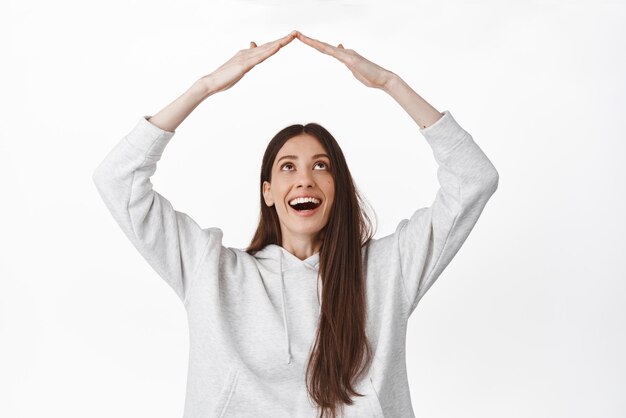 Family and insurance concept Happy smiling woman looking at her hands making rooftop home roof gesture standing upbeat against white background