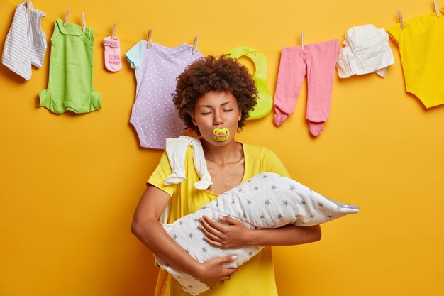 Family, motherhood and parenting concept. Mother holds small baby on hands, enjoys moment of being mum, poses with nipple in mouth and closed eyes, rope with newborn clothes behind, yellow wall