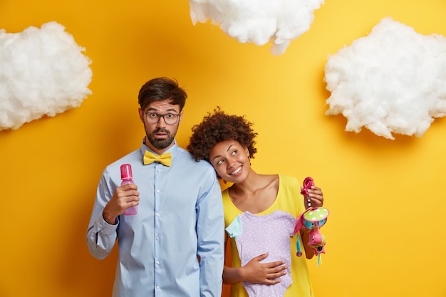 Free photo family, parenthood concept. surprised future father holds feeding bottle, dreamy pregnant wife leans at shoulder of husband, holds bodysuit and mobile, imagines their new happy life with baby