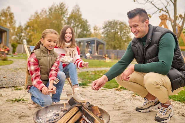 Family, picnic. Young adult caucasian smiling man pretty woman and girl school age crouched near bonfire with marshmallows on stick in yard of house on autumn day