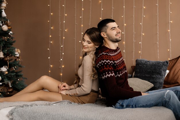 Family portrait. Man and woman relax on soft grey bad in a room with Christmas tree