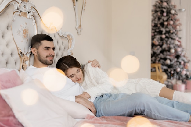 Family portrait. Man and woman relax on soft grey bad in a room with Christmas tree