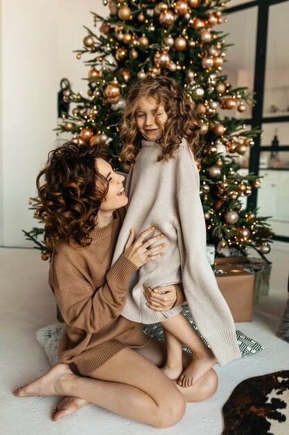 크리스마스 트리 앞에서 포즈를 취하는 니트 옷을 입은 작은 딸과 함께 젊은 매력적인 어머니의 가족 초상화