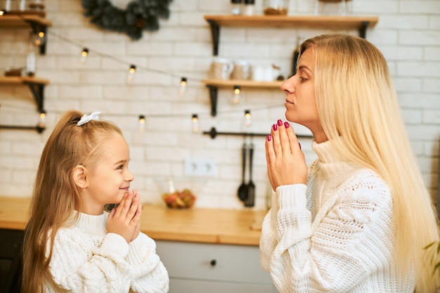 Family and relationships concept. Charming young blonde mom teaching her little daughter to perform tricks sitting at kitchen counter with hands pressed, having cakes and coffee for dessert