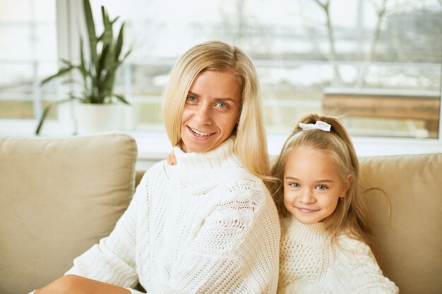 Family, relationships, generation, love and bonding concept. Stylish young European mom with straight long hair smiling relaxing on comfortable sofa, sitting next to her adorable daughter