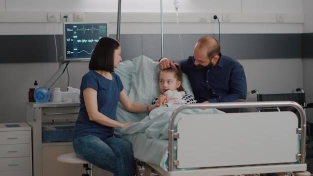 Family sitting beside hospitalized sick daughter discussing medication recovery treatment during medicine examination in hospital ward. Child resting in bed after suffering disease infection surgery