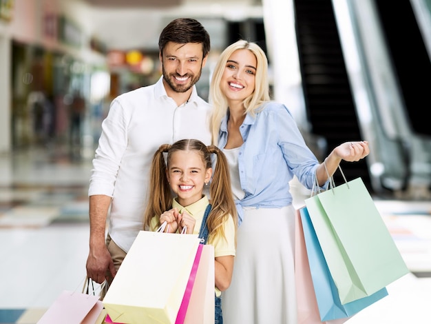 Family of three posing with shopping bags smiling at camera standing in mall center. seasonal sales concept