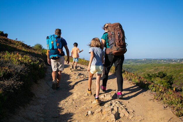 トラックを歩いているバックパックを持つ旅行者の家族。両親と2人の子供が屋外でハイキングします。背面図。アクティブなライフスタイルや冒険旅行のコンセプト