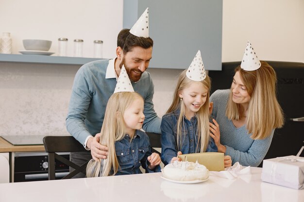 Family and two their daughters celebrate birthday in the kitchen. People wear a party-hat. Girl keep a boxes with presents.