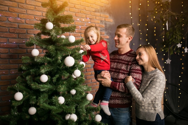 小さな娘がおもちゃをクリスマスツリーに吊るしている家族