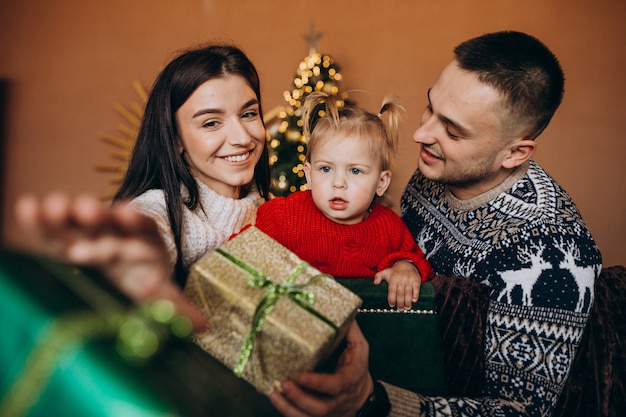 Famiglia con la piccola figlia che si siede dall'albero di natale e che disimballa il contenitore di regalo