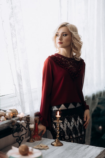 Fancy blonde woman dressed in red clothes stands in a room with Christmas decor