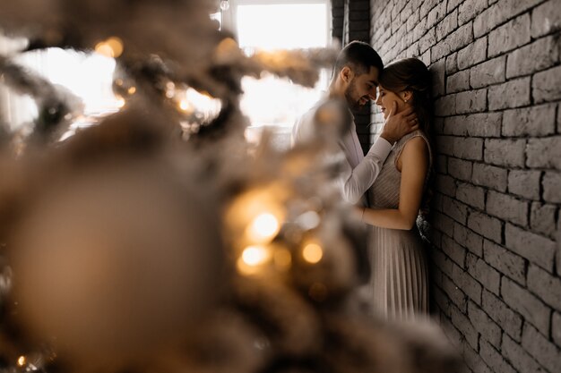 Fancy dressed man and woman in silver gown hug each other tender standing before a Christmas tree