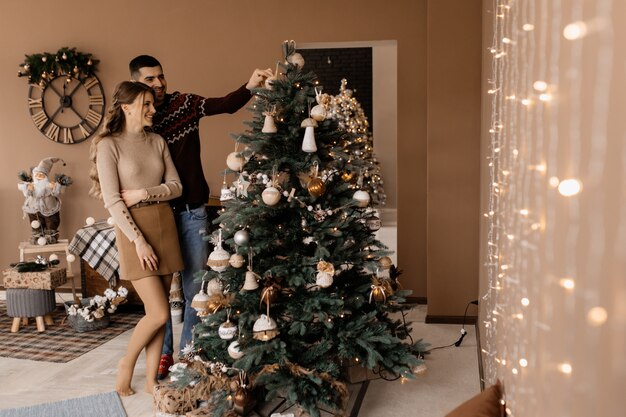 Fancy dressed man and woman in silver gown hug each other tender standing before a Christmas tree