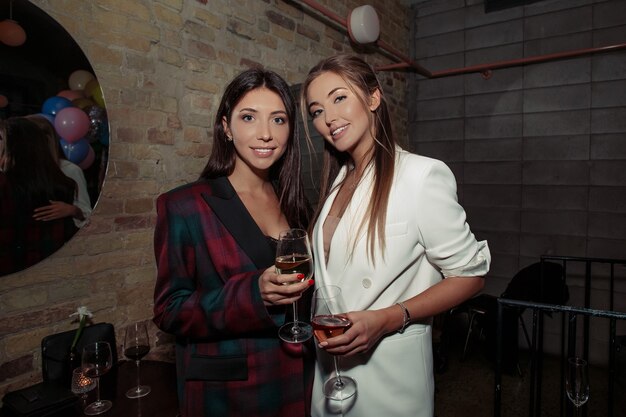 Fancy girls having fun on a party. Blonde wearing white jacket and accessories, natural makeup on the face and stylish hair. Brunette woman in a black fashionable dress.