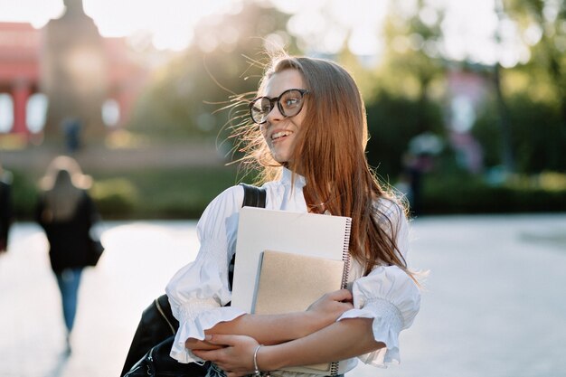 Fantastic happy young female student back to college with notebooks and laptop, going to university with happy smile on sunny street