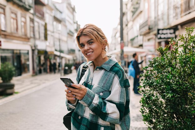 ヨーロッパの街を歩きながらスマートフォンを使用して明るいシャツを着ているブロンドの髪の素晴らしいきれいな女性秋にヨーロッパを旅行している女の子