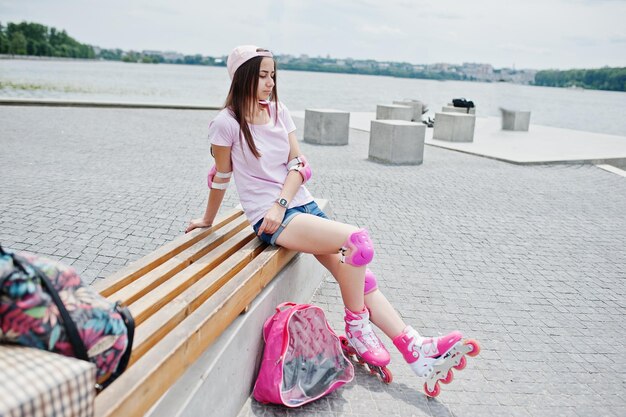 Fantastic young woman in casual clothing and cap sitting on the bench in the skatepark with rollerblades on