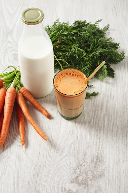 Raccolto di carote dell'azienda agricola che si trova vicino alla bottiglia per il latte e al bicchiere riempito con succo fresco naturale e latte con cannuccia dorata