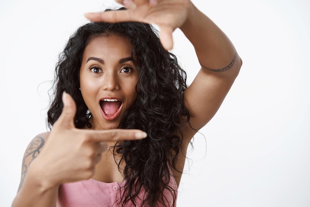 Affascinata ragazza afroamericana che controlla cosa incredibile attraente donna tatuata dai capelli ricci che fa cornici a mano e ansimante sguardo sorpreso stupito fotocamera sfondo bianco