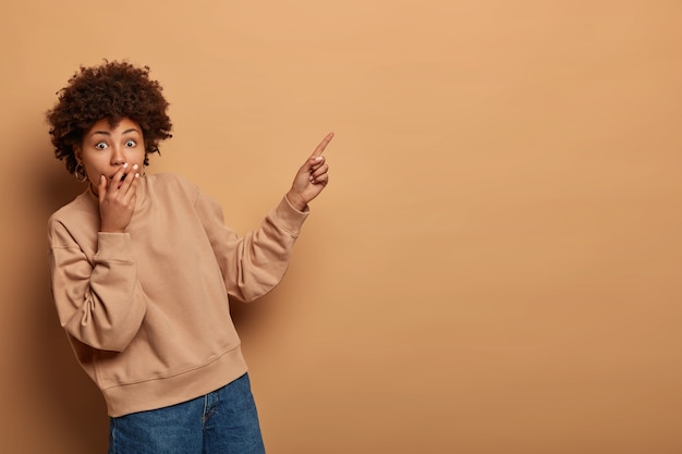 Free photo fascinated curly haired woman covers mouth with shock, points index finger at upper right corner, demonstrates astonishng product