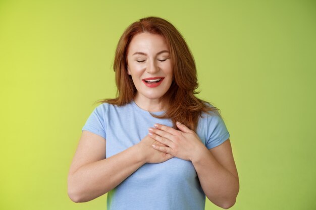 Fascinated cute redhead passionate middleaged woman sighing lovely touch heart close eyes smiling delighted express admiration temptation feeling appreciation grateful emotions green wall