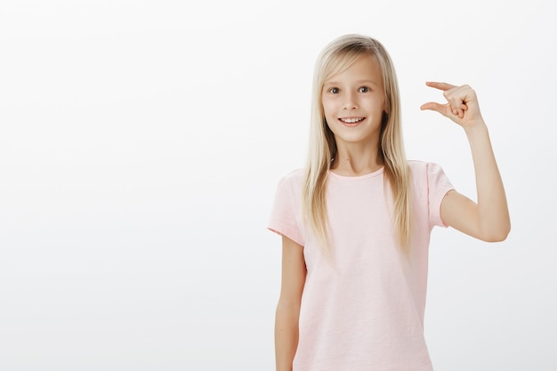 fascinated dreamy little girl with blond hair in pink t-shirt, shaping something small or tiny with fingers, amazed with little bird she saw in park, standing surprised over gray wall