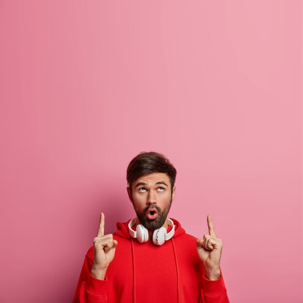 Очарованный, впечатленный бородатый мужчина, меломан показывает вверху, показывает вверх что-то удивительное, использует стереонаушники для прослушивания музыки, задыхается от удивления, изолирован на розовой стене. Концепция промо