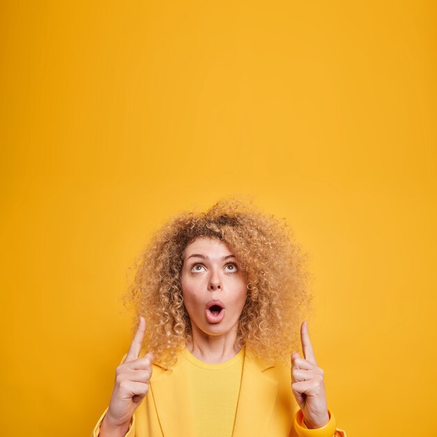 Fascinated surprised curly haired European woman points above impressed by awesome promo keeps mouth opened dressed elegantly isolated over yellow wall shows interesting shopping deal.