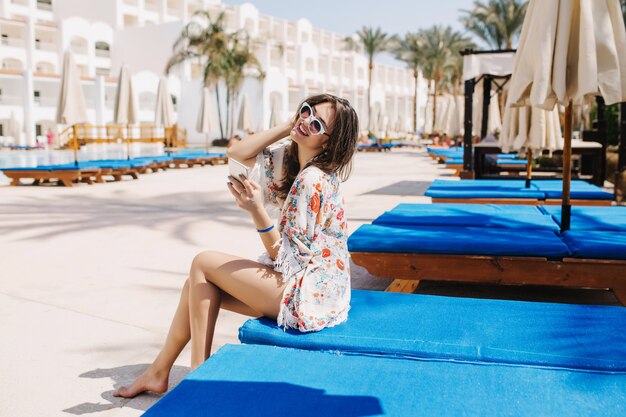 Fascinating barefooted dark-haired girl having fun outside, resting on chaise-longue near the hotel. Portrait of cute tanned young woman in sunglasses and trendy attire sitting on trestle-bed