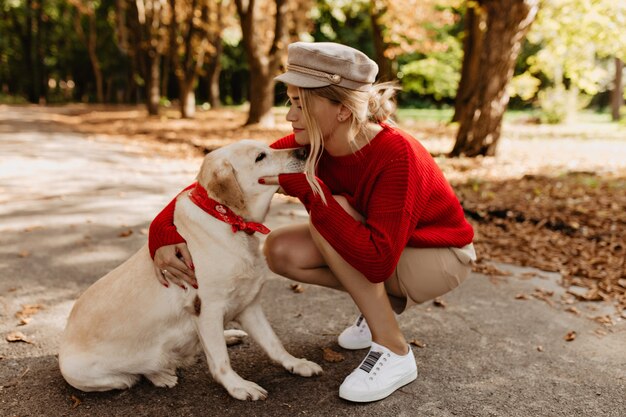 秋の公園で一緒に一日を過ごす愛らしいラブラドールと魅力的なブロンド。彼女の最愛の犬を抱き締める季節の服を着た女の子の感動的な写真。