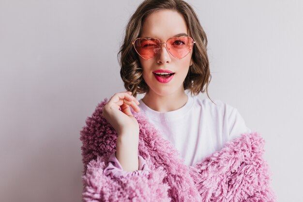 Fascinating brunette girl in pink fur coat playfully posing on white wall. lovable graceful woman in fluffy jacket.